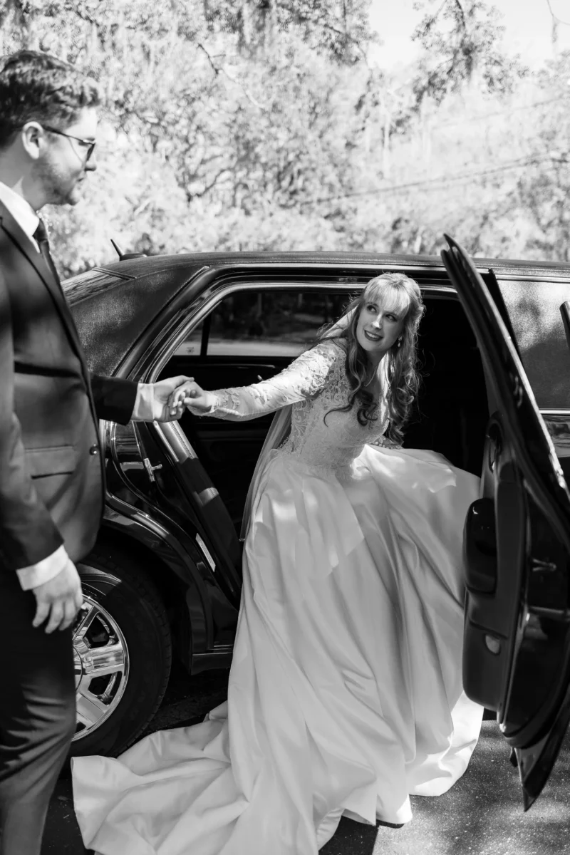 A bride in a flowing white gown steps out of a black limousine, assisted by a man in a suit. Sunlight filters through the trees. A bride in a flowing white gown steps out of a black limousine, assisted by a man in a suit. Sunlight filters through the trees.