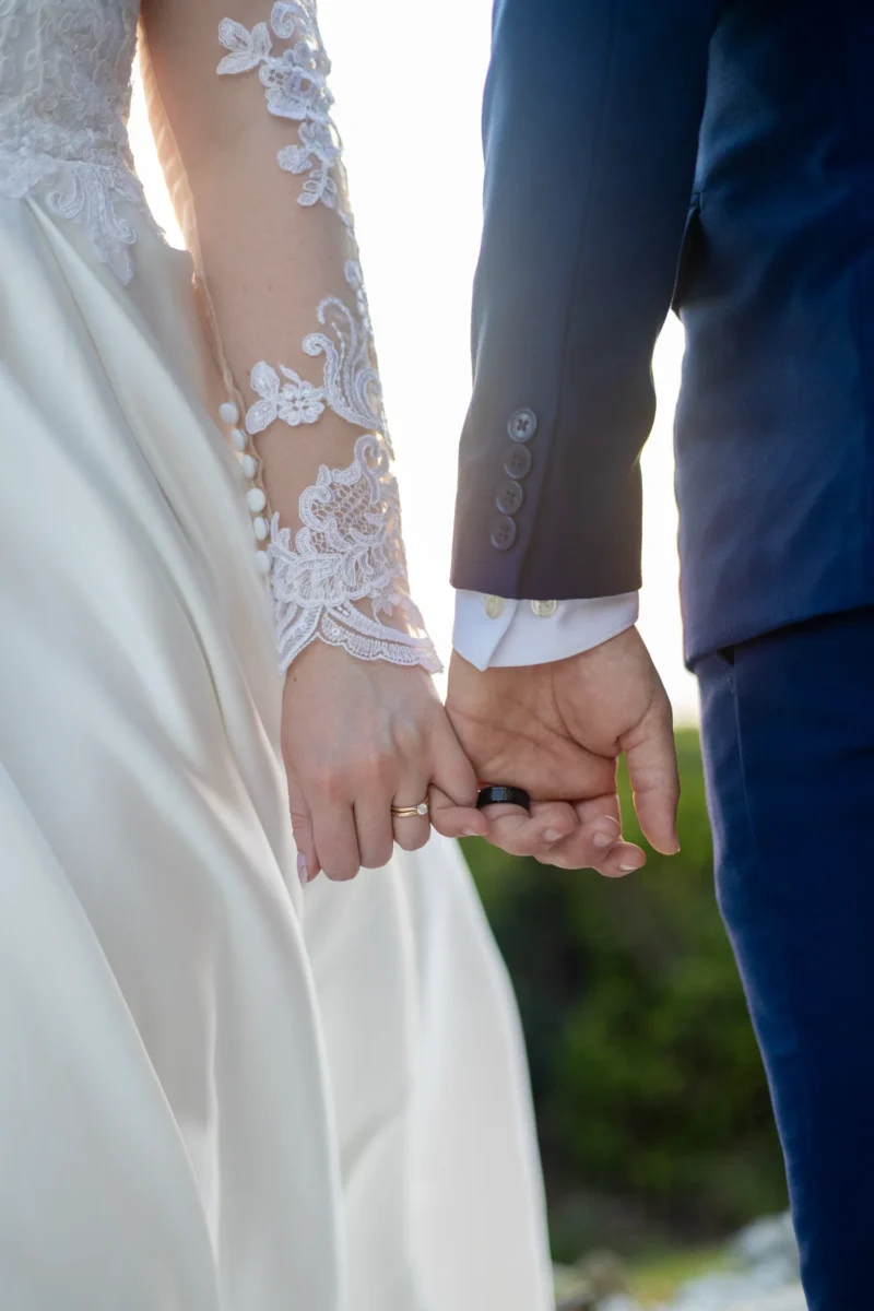 A close-up of a bride and groom holding hands, showcasing their wedding bands and elegant attire against a softly lit background. A close-up of a bride and groom holding hands, showcasing their wedding bands and elegant attire against a softly lit background.