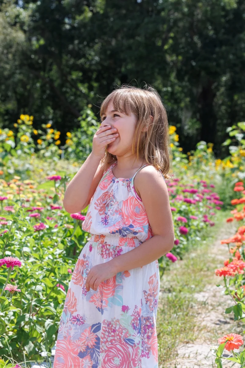A young girl stands amidst vibrant flowers, wearing a colorful dress and touching her cheek, enjoying a sunny day in a garden. A young girl stands amidst vibrant flowers, wearing a colorful dress and touching her cheek, enjoying a sunny day in a garden.