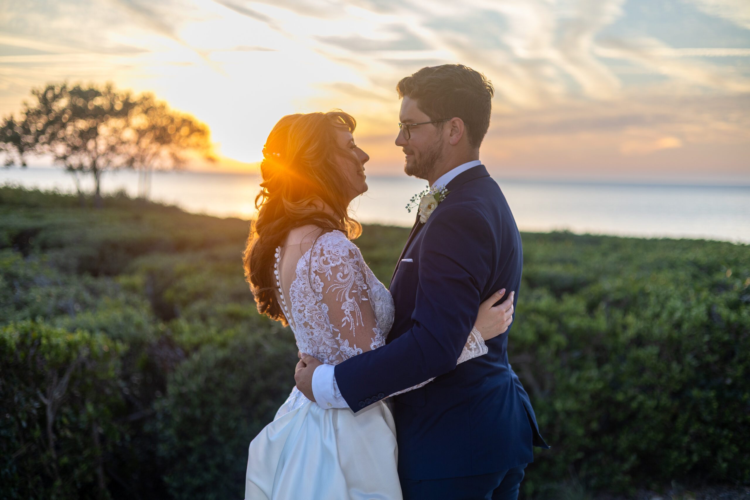 LO1A1530 A pregnant woman in a white lace dress cradles her baby bump while holding a vibrant sunflower surrounded by greenery.